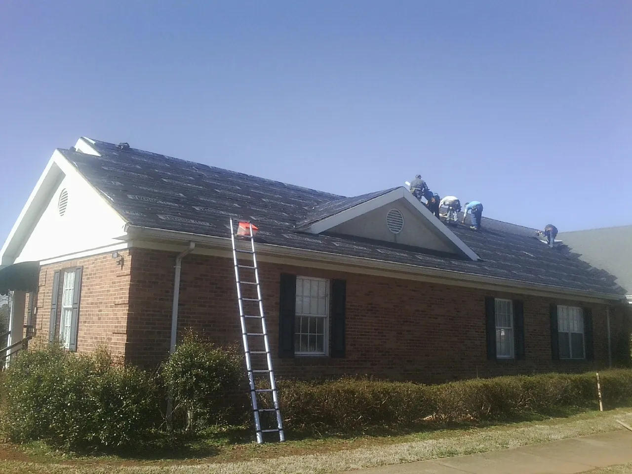 Crew tearing shingles off a brick ranch roof with a ladder set — Metal Roof Repair work by our crew in Flat Rock